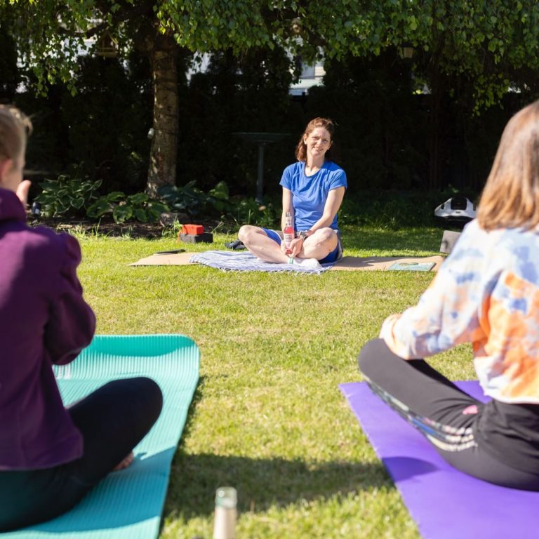 Frau meditiert im Freien, umgeben von zwei entspannenden Yoga-Teilnehmern.