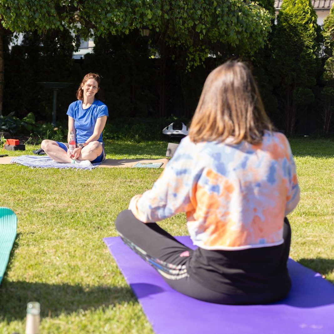 Zwei Frauen beim Yoga im Freien auf Matten im grassbewachsenen Garten.